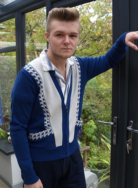 Man wearing a blue sweater with a patterned design, standing indoors with greenery outside.