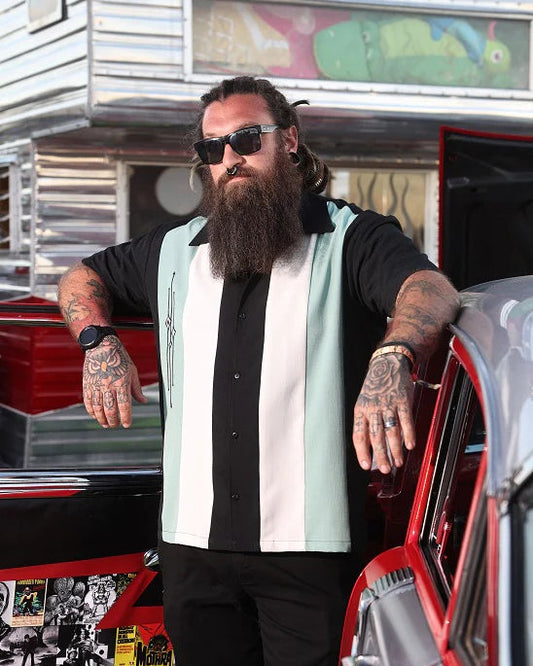 Man with tattoos and sunglasses standing next to a vintage trailer and car.