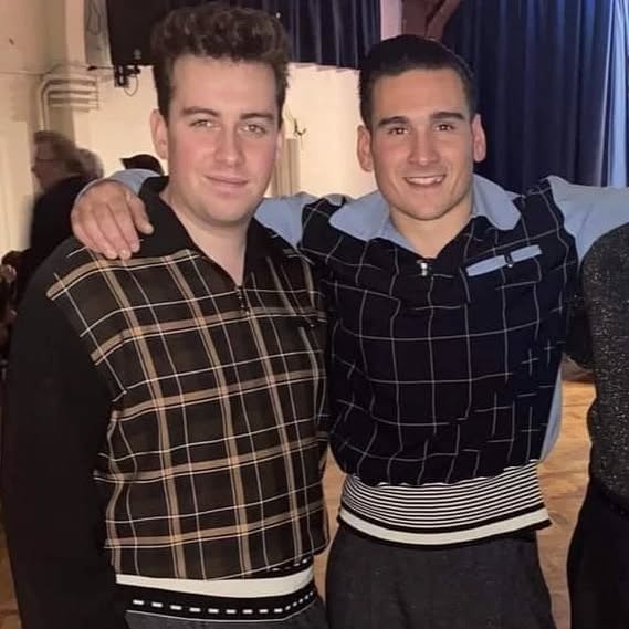 Two men posing together wearing matching checkered outfits indoors.