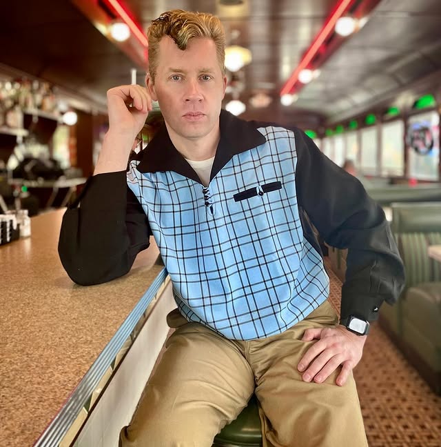 Man sitting at a bar counter wearing a blue plaid shirt and beige pants.