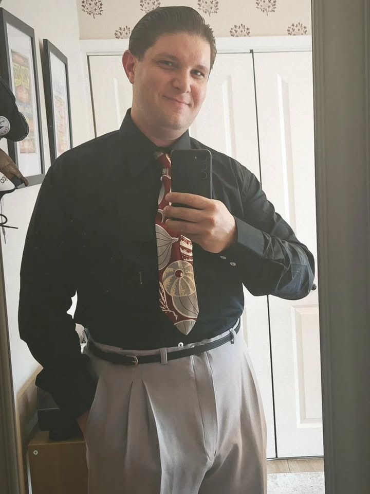 Person wearing a black shirt, patterned tie, and light-colored pants in front of a mirror.