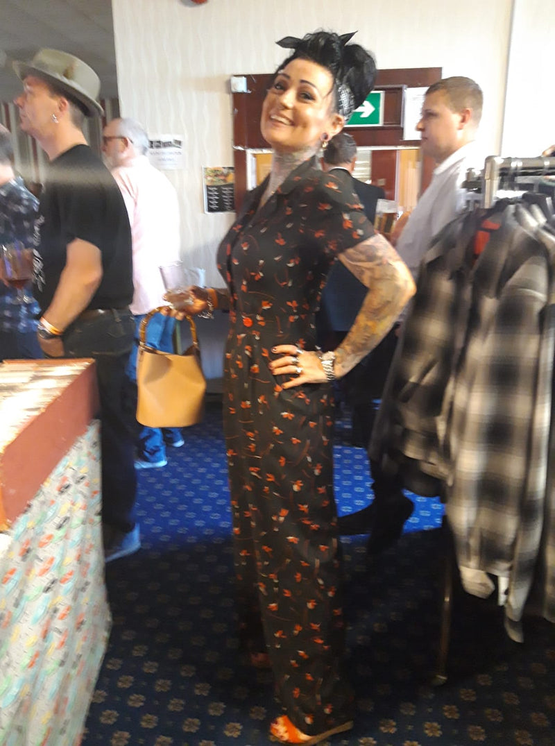 Woman in a patterned jumpsuit standing in a room with people and clothing around